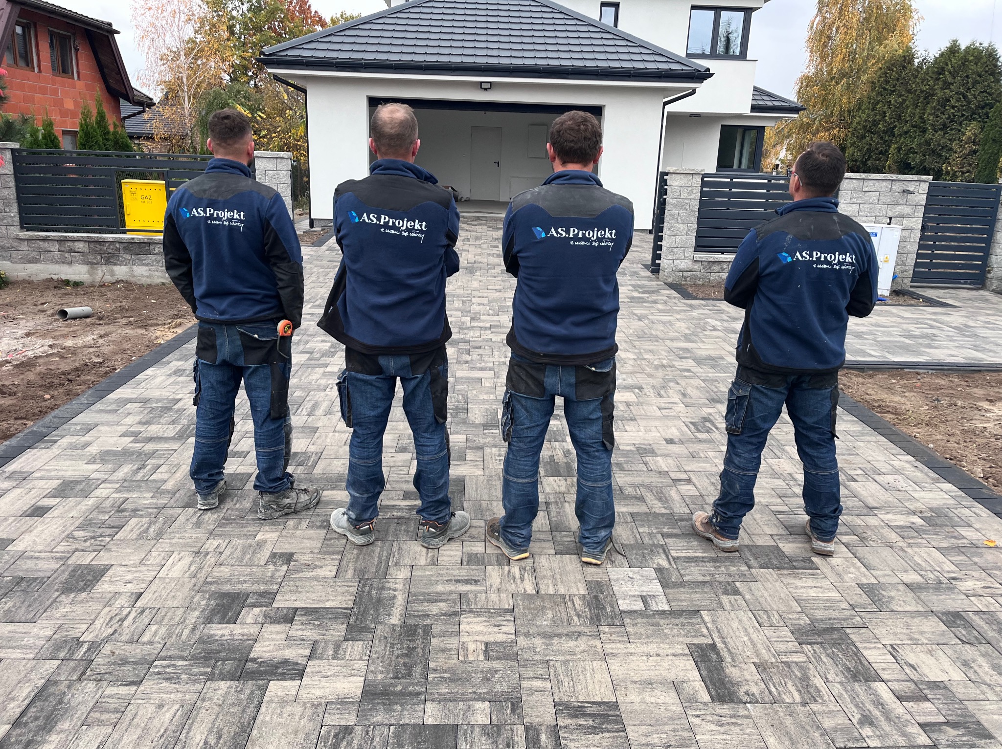 Ułożona kostka brukowa w odcieniach szarości przed nowoczesnym domem; czwórka pracowników firmy w granatowych bluzach z logo stoi tyłem, obserwując efekt końcowy.