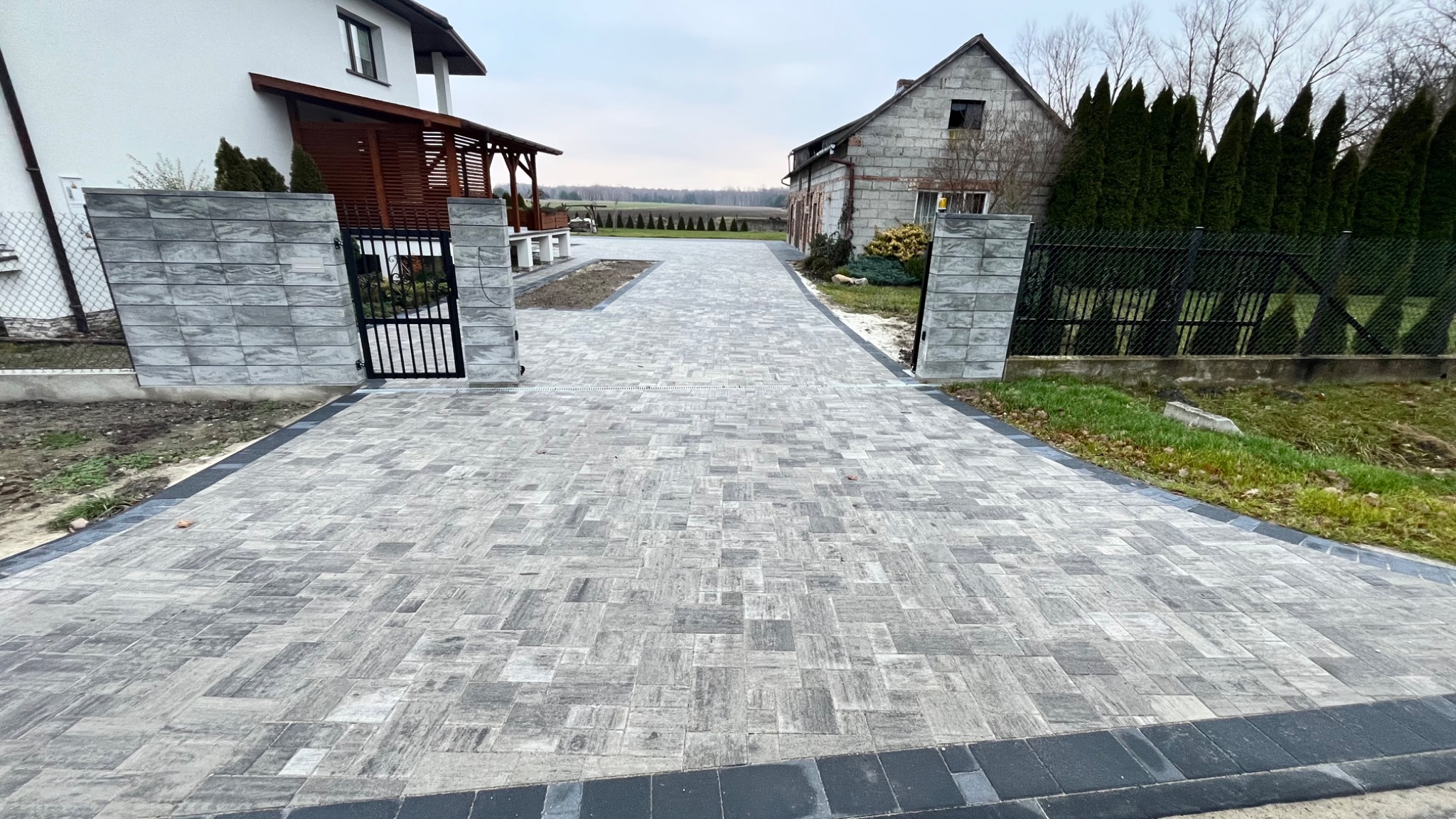 Szeroki, szary podjazd z kostki brukowej prowadzący pomiędzy dwoma domami, obrzeża wykończone czarną kostką.