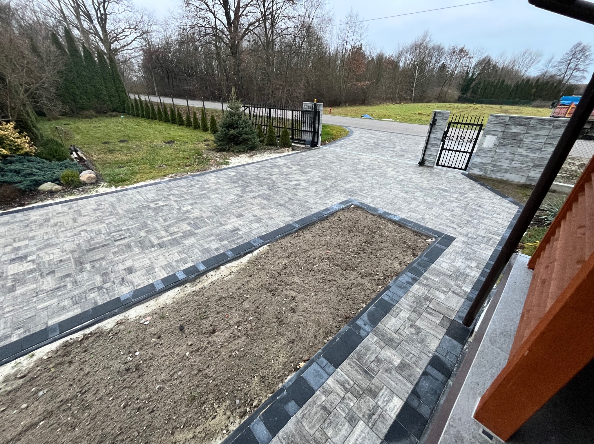 Ułożona szara kostka brukowa z ciemnoszarą obwódką, tworząca ścieżkę i podjazd do posesji z bramą w tle, widok z góry.