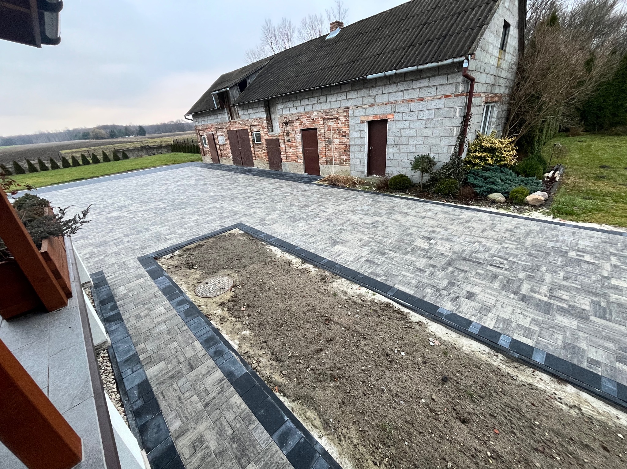 Szeroki, wybrukowany szarym kamieniem podjazd prowadzący do budynku gospodarczego z cegły, z wyznaczonym miejscem na rabatę w trakcie przygotowania, widok z góry.