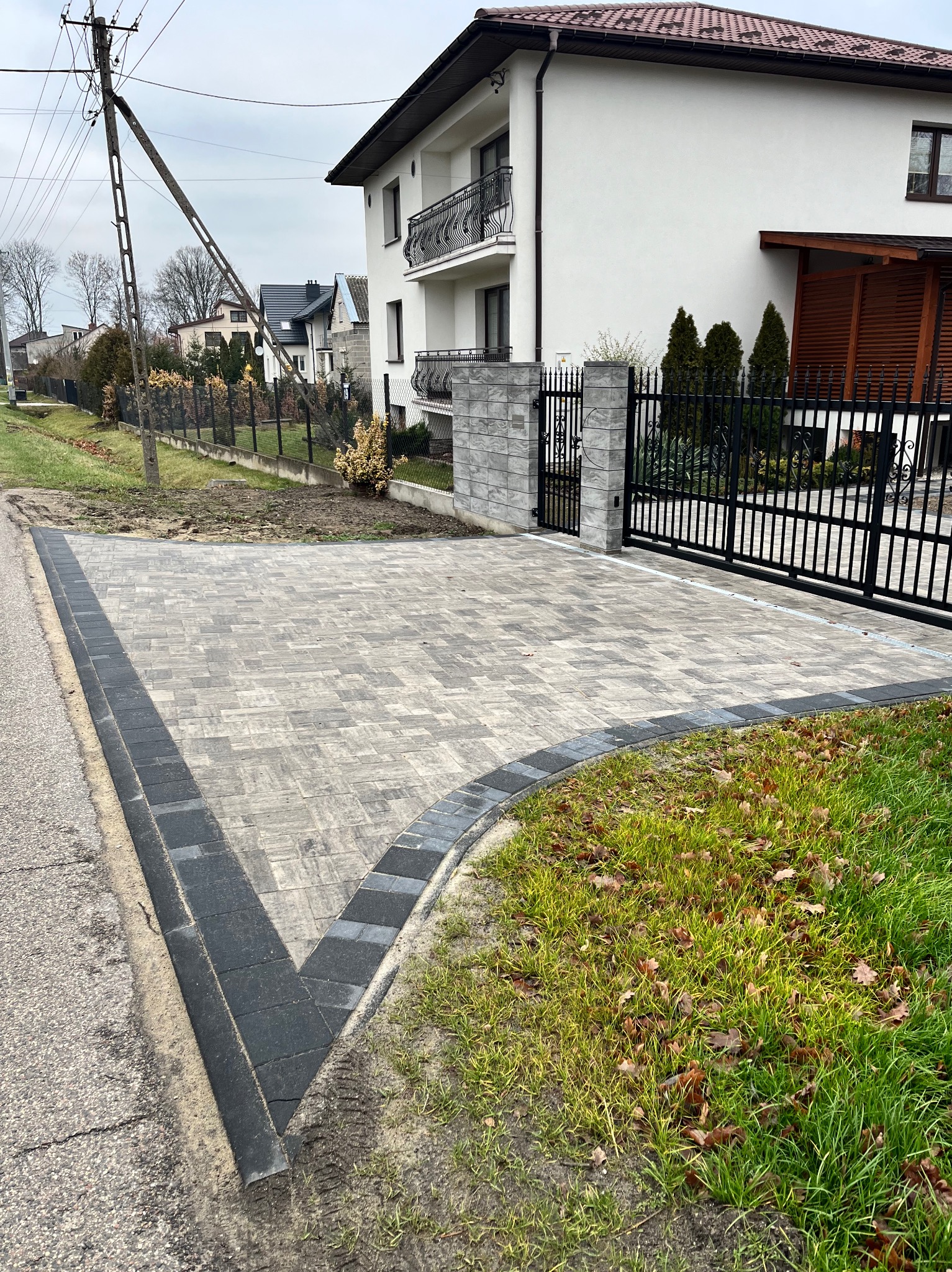 Ułożona szara kostka brukowa z ciemnoszarą obwódką na podjeździe przed nowoczesnym domem z czarną bramą i ogrodzeniem, widoczne ślady piasku na obrzeżach.