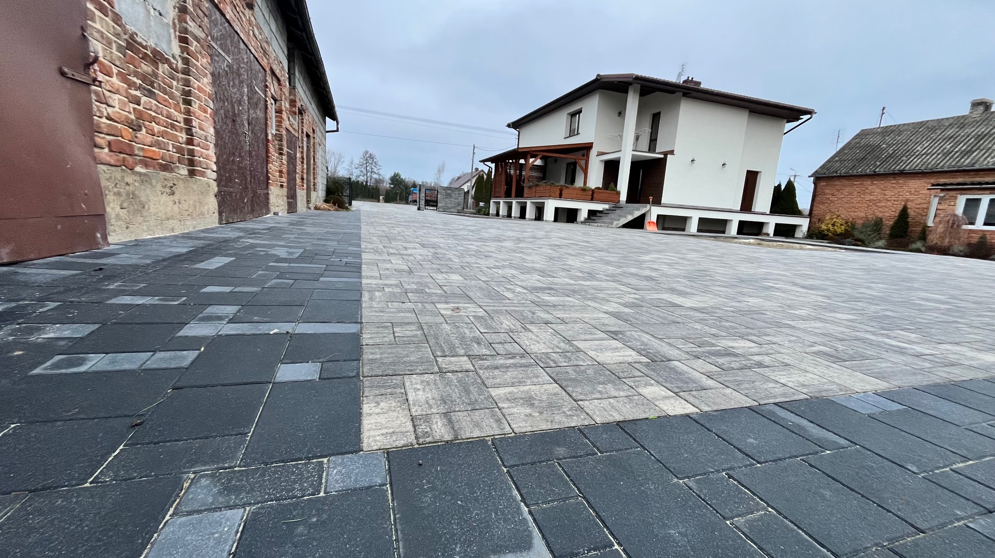 Szeroki podjazd z szarej kostki brukowej ułożonej we wzór prostokątów, obrzeża z ciemnej kostki, w tle budynki mieszkalne i pochmurne niebo.