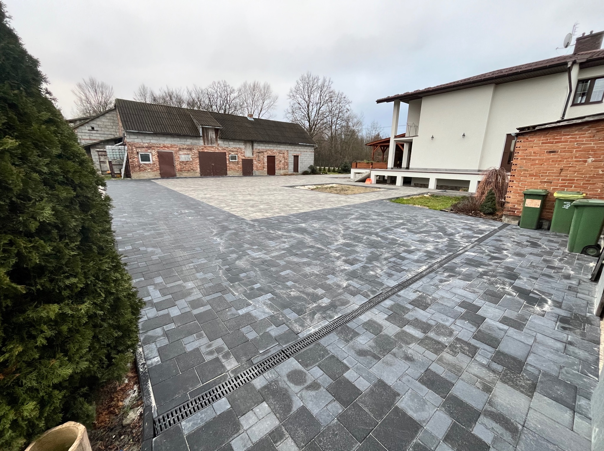 Nowo ułożona, szara kostka brukowa na podjeździe, z widocznym odpływem liniowym, domem z białym tynkiem i budynkiem gospodarczym w tle, w pochmurny dzień.