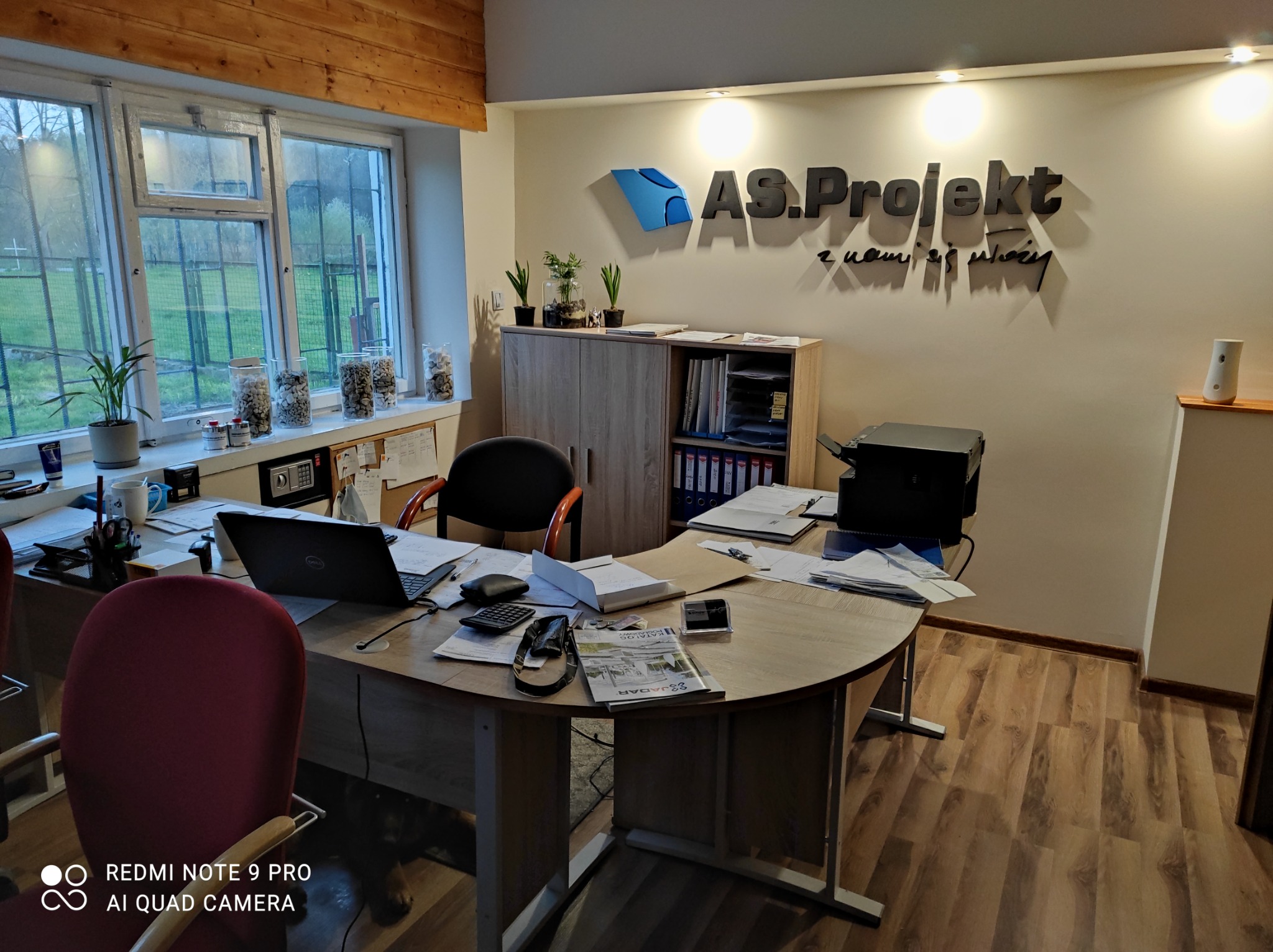 Biuro firmy projektowej z drewnianą podłogą, biurkiem z laptopem i katalogiem firmy SSJADAR, widok na okno i logo firmy AS.Projekt na ścianie.