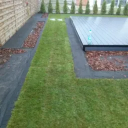 Nowo założony trawnik z wytyczoną ścieżką i podwyższonym tarasem z kompozytu, obok drewnianego ogrodzenia i rzędu małych iglaków.