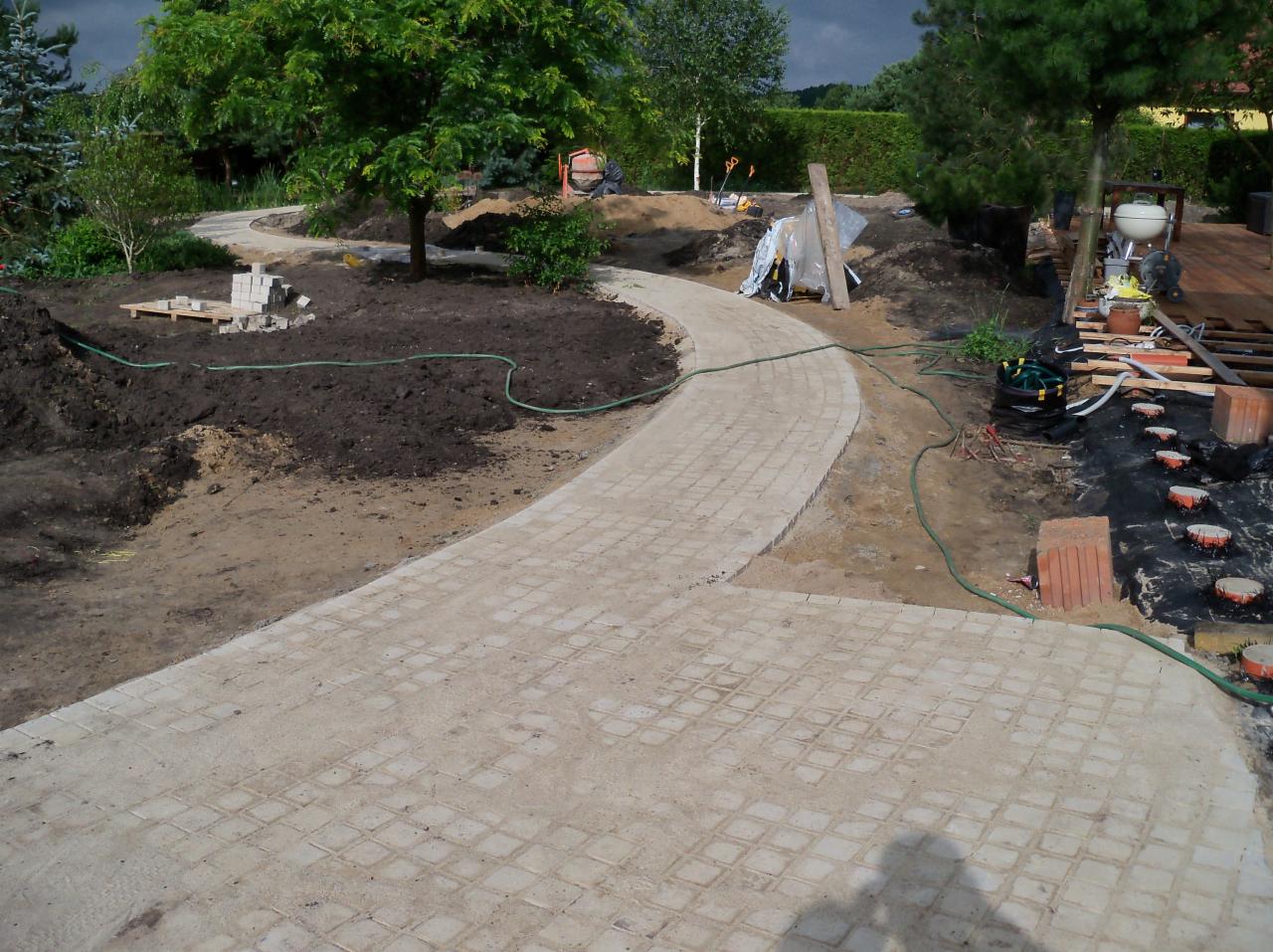Krzywa ścieżka z jasnej kostki brukowej w trakcie budowy, widoczne przygotowane podłoże, materiały budowlane i elementy ogrodu w tle.