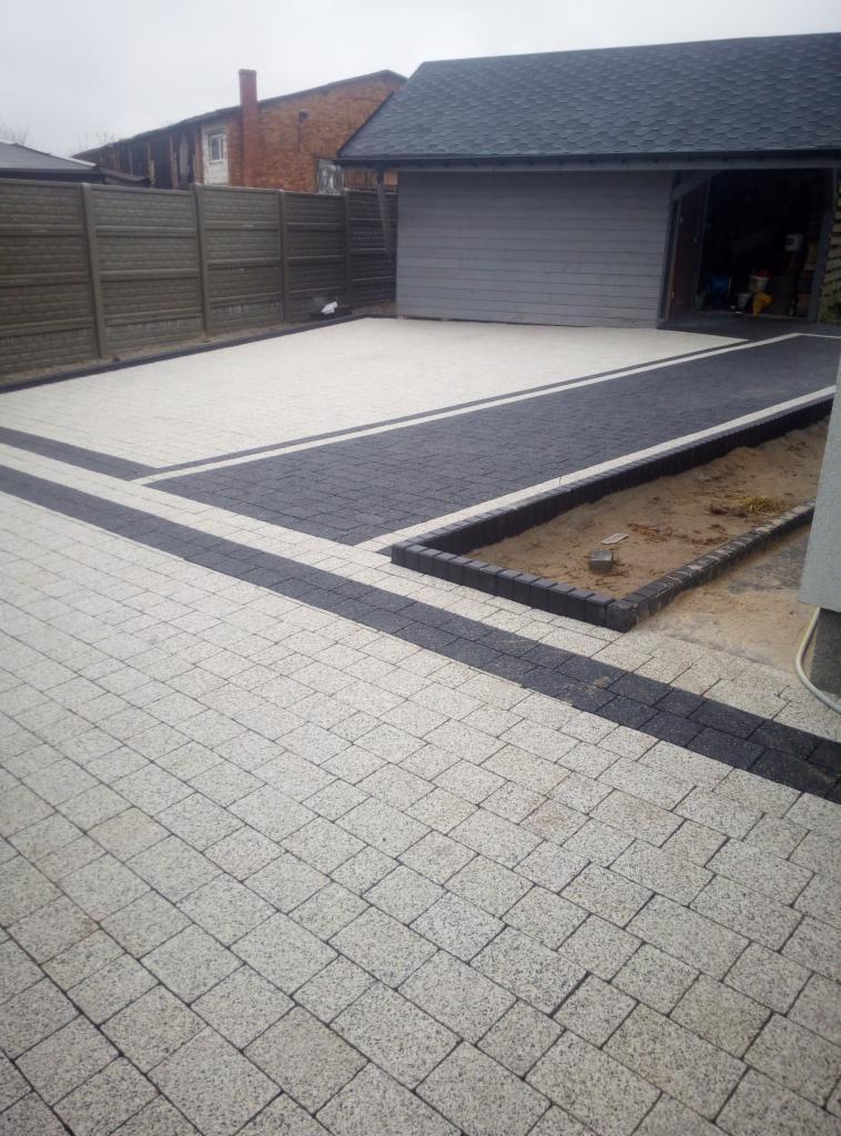 Ułożona jasnoszara kostka brukowa z ciemnymi pasami tworzącymi wzór na podjeździe, obok garażu i ogrodzenia panelowego.