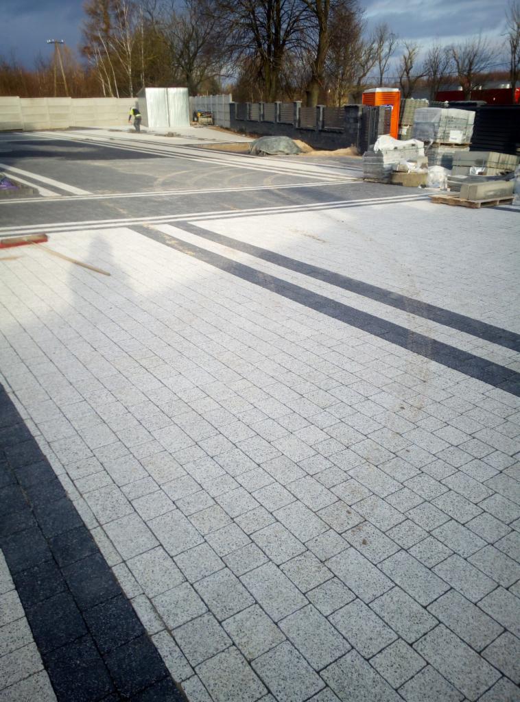 Ułożona jasnoszara kostka brukowa z ciemnymi pasami, widoczny pracownik w oddali i materiały budowlane.