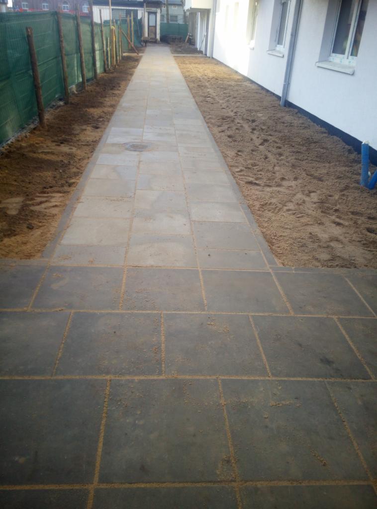 Długa ścieżka z szarych płyt chodnikowych prowadząca wzdłuż budynku z białymi ścianami, z widocznymi świeżo przygotowanymi rabatami po obu stronach.