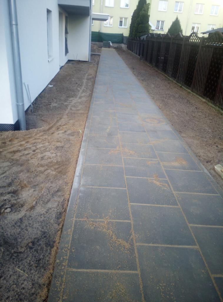 Długa, prostokątna ścieżka z szarych płyt betonowych, wypełnionych piaskiem, prowadząca wzdłuż budynku i ogrodzenia z siatki, z widoczną ziemią przygotowaną pod trawnik.