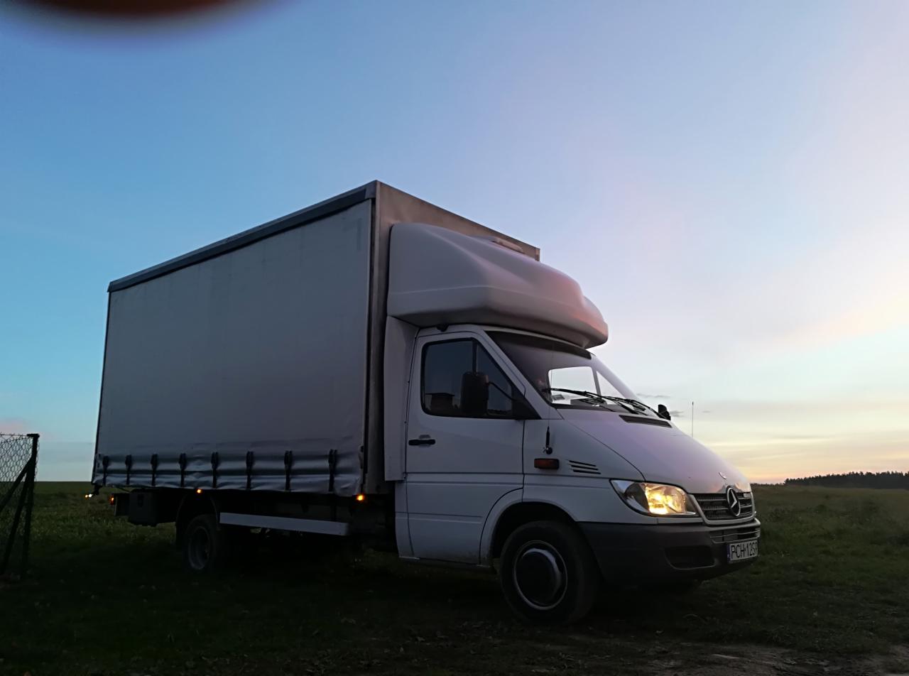 Biały samochód dostawczy typu bus z plandeką, zaparkowany na trawiastym terenie o zmierzchu, widoczne włączone światła mijania i tablica rejestracyjna.