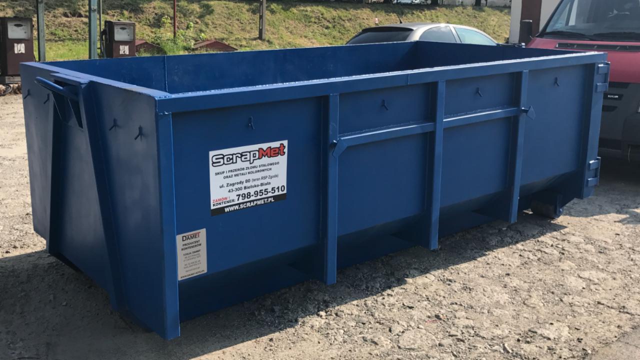 Duży, niebieski kontener na złom z naklejkami firm ScrapMet i Damet na tle stacji benzynowej i zaparkowanych aut.