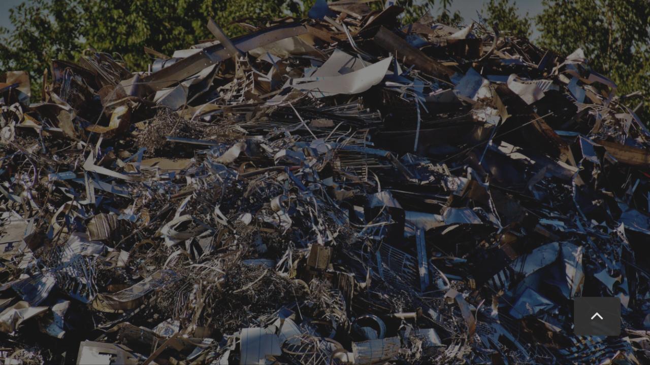 Góra złomu metalowego, w tym blachy, drutu i elementów konstrukcyjnych, składowana na zewnątrz na tle zieleni drzew.