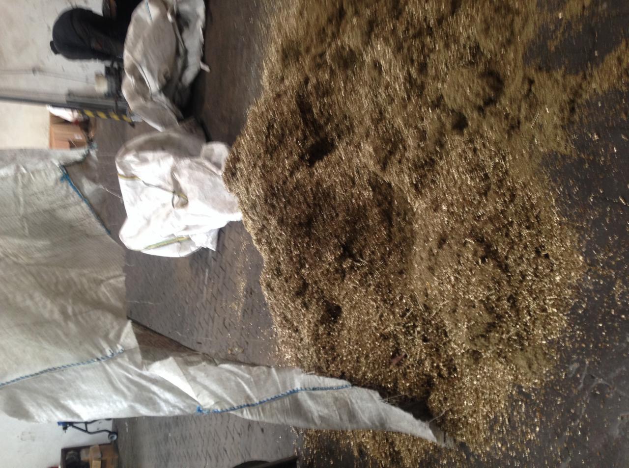 Sterta drobnych, metalowych wiórów w odcieniach złota i brązu, wysypana obok worków na metalowej, wzorzystej podłodze w hali.