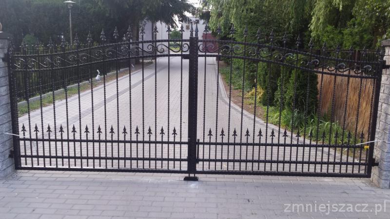 Czarna, metalowa brama wjazdowa z ozdobnymi elementami, prowadząca do posesji z brukowaną drogą, otoczona zielenią.