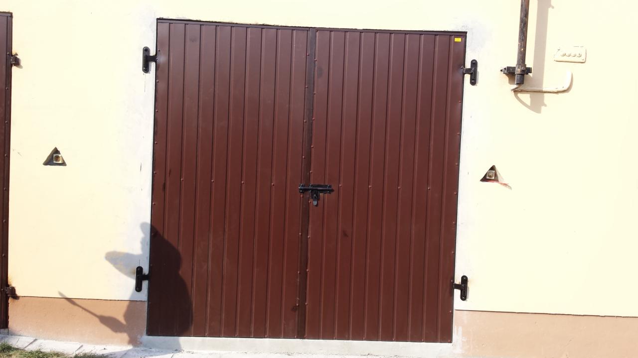 Brązowa, metalowa brama garażowa z pionowymi panelami i czarnym zamkiem na tle jasnej ściany budynku z widocznymi instalacjami.