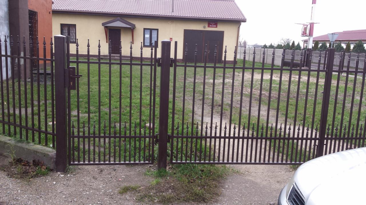 Metalowa brama wjazdowa i ogrodzenie posesji z elementami dekoracyjnymi w kolorze ciemnego brązu, z widocznym zamkiem i fragmentem białego samochodu w prawym dolnym rogu.