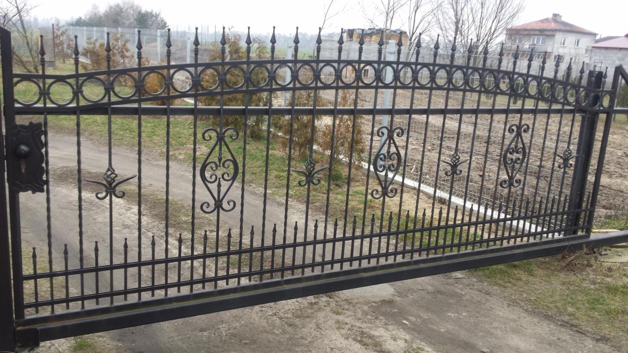 Czarna, metalowa brama wjazdowa z ozdobnymi elementami, zamknięta na zamek, widok na posesję z domem w tle.