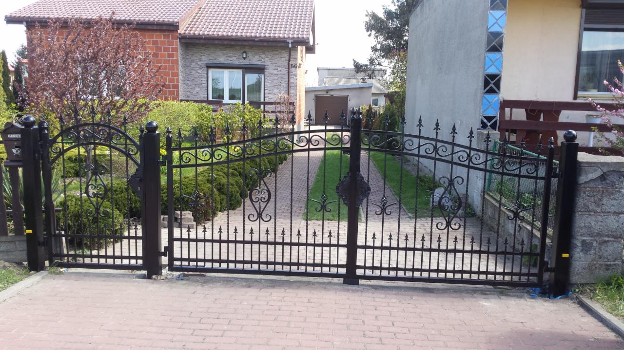 Kuta, metalowa brama wjazdowa w kolorze czarnym z dekoracyjnymi elementami, prowadząca do domu z cegły i garażu w tle, widoczna skrzynka na listy.