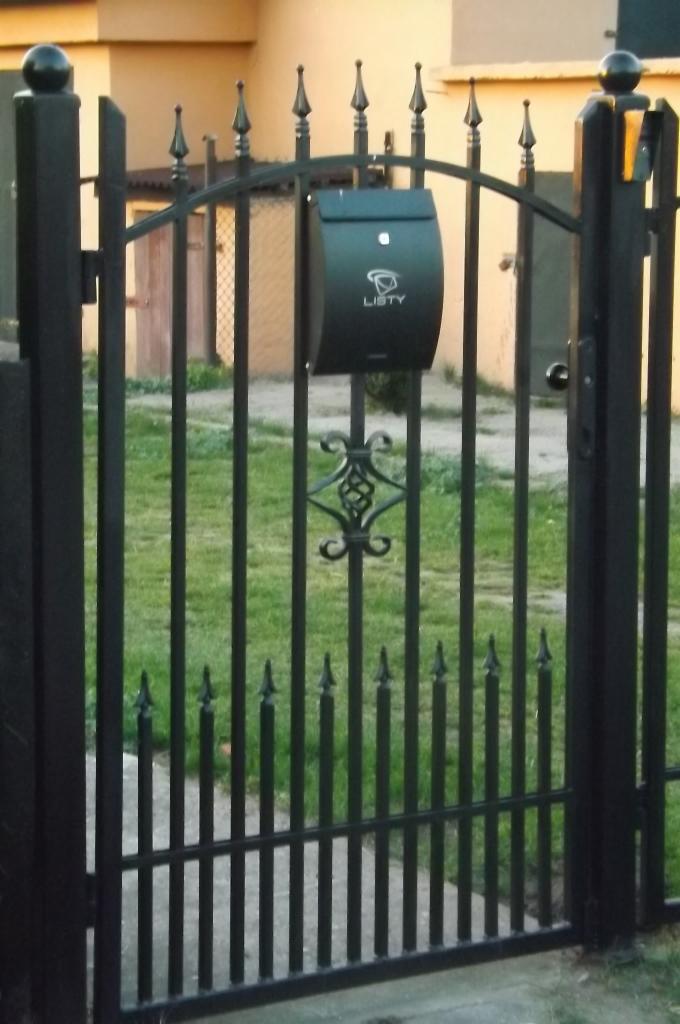 Czarna, metalowa brama z dekoracyjnymi elementami, zakończona ostrymi grotami, z zamontowaną skrzynką na listy z napisem 'LISTY' i ozdobnym motywem roślinnym pośrodku.