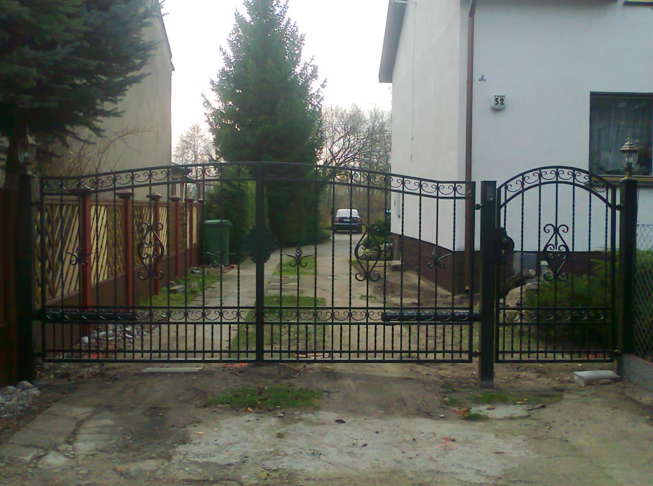 Czarna, metalowa brama wjazdowa z ozdobnymi elementami, prowadząca do posesji z widocznym budynkiem z numerem 52 i samochodem w tle.