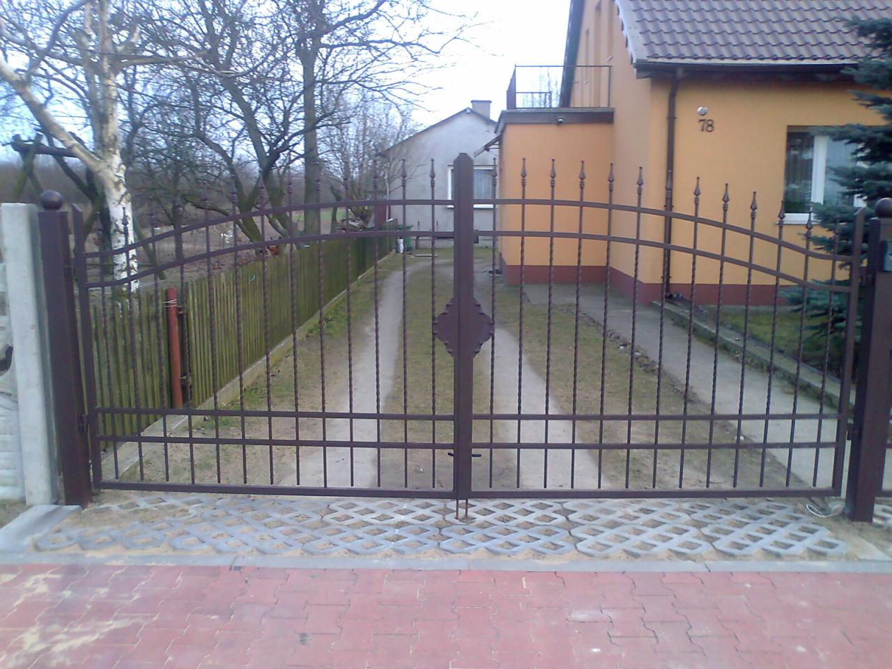 Nowa, brązowa, metalowa brama wjazdowa z dekoracyjnym elementem rombu na środku, osadzona na betonowych słupkach i kostce brukowej, prowadząca do domu z numerem 78.
