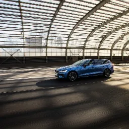 Niebieski samochód kombi Volvo V60 porusza się w tunelu z metalową konstrukcją dachową, tworzącą geometryczne cienie na asfalcie. W tle widoczny drugi pojazd.