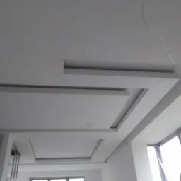 Biały sufit podwieszany z geometrycznymi wgłębieniami i wystającymi elementami, widoczne okno i fragment pomieszczenia w trakcie prac wykończeniowych.