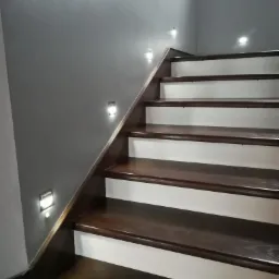 Schody z ciemnego drewna z białymi podstopnicami oświetlone małymi, kwadratowymi lampami ściennymi LED, zamontowanymi na szarej ścianie wzdłuż biegu schodów.