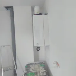 Biały piec grzewczy wiszący na ścianie w trakcie montażu, widoczne przewody, drabina i zabezpieczony folią karton w pomieszczeniu, w tle białe ściany i rura wentylacyjna.