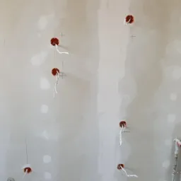 Ściana w trakcie remontu z widocznymi puszkami elektrycznymi z wystającymi przewodami i zaznaczonymi punktami pomiarowymi, oraz fragment instalacji hydraulicznej w dolnym rogu.