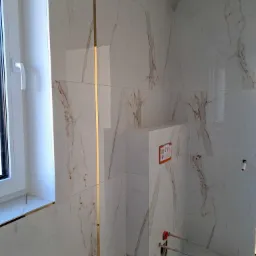 Ściany i podłoga wyłożone białymi płytkami imitującymi marmur z brązowymi żyłami, widoczna złota listwa dekoracyjna, stelaż podtynkowy WC z przyłączami wodnymi, okno z białymi ramami.