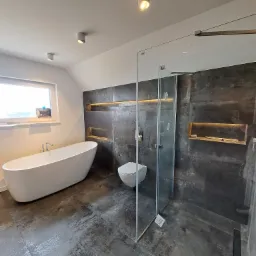 Nowoczesna łazienka na poddaszu z wolnostojącą wanną, szarą płytką imitującą beton, kabiną prysznicową typu walk-in i podświetlanymi niszami w ścianie.