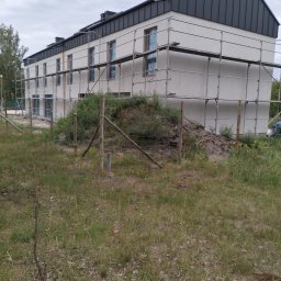 RGB Axbud - Budynek w trakcie ocieplania elewacji, widoczne rusztowania i częściowo otynkowana ściana, ogrodzenie z drewnianych słupków i siatki, zaniedbany teren wokół.