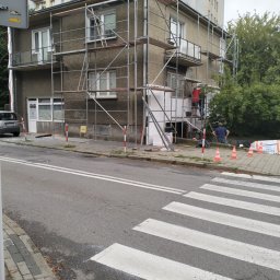 RGB Axbud - Dwupiętrowy budynek w trakcie prac elewacyjnych, z widocznym rusztowaniem, pracownikami na rusztowaniu oraz stosami białego styropianu na chodniku, na tle bloku mieszkalnego i przejścia dla pieszych.
