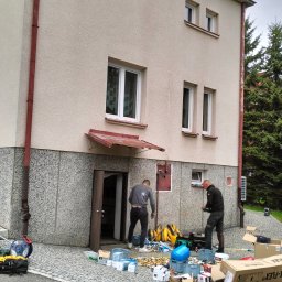 RGB Axbud - Dwóch mężczyzn przy budynku z elewacją, rozpakowujących materiały hydrauliczne na chodniku z kostki brukowej, widoczne skrzynki, rury i narzędzia.