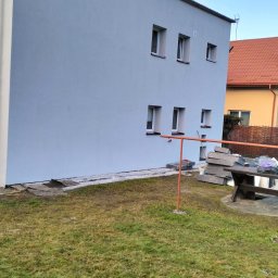 RGB Axbud - Świeżo pomalowana ściana budynku z widocznymi oknami, czerwonymi kominami i fragmentem trawnika z elementami budowlanymi w tle.