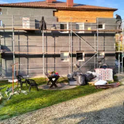 Dom w trakcie ocieplania styropianem, widoczne rusztowania, przygotowane materiały budowlane, narzędzia na stole i pracownicy na rusztowaniu, częściowo ocieplona ściana budynku.