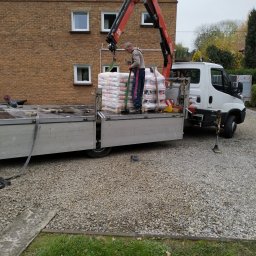 RGB Axbud - Rozładunek worków z zaprawą elewacyjną za pomocą dźwigu samochodowego na tle ceglanego budynku.