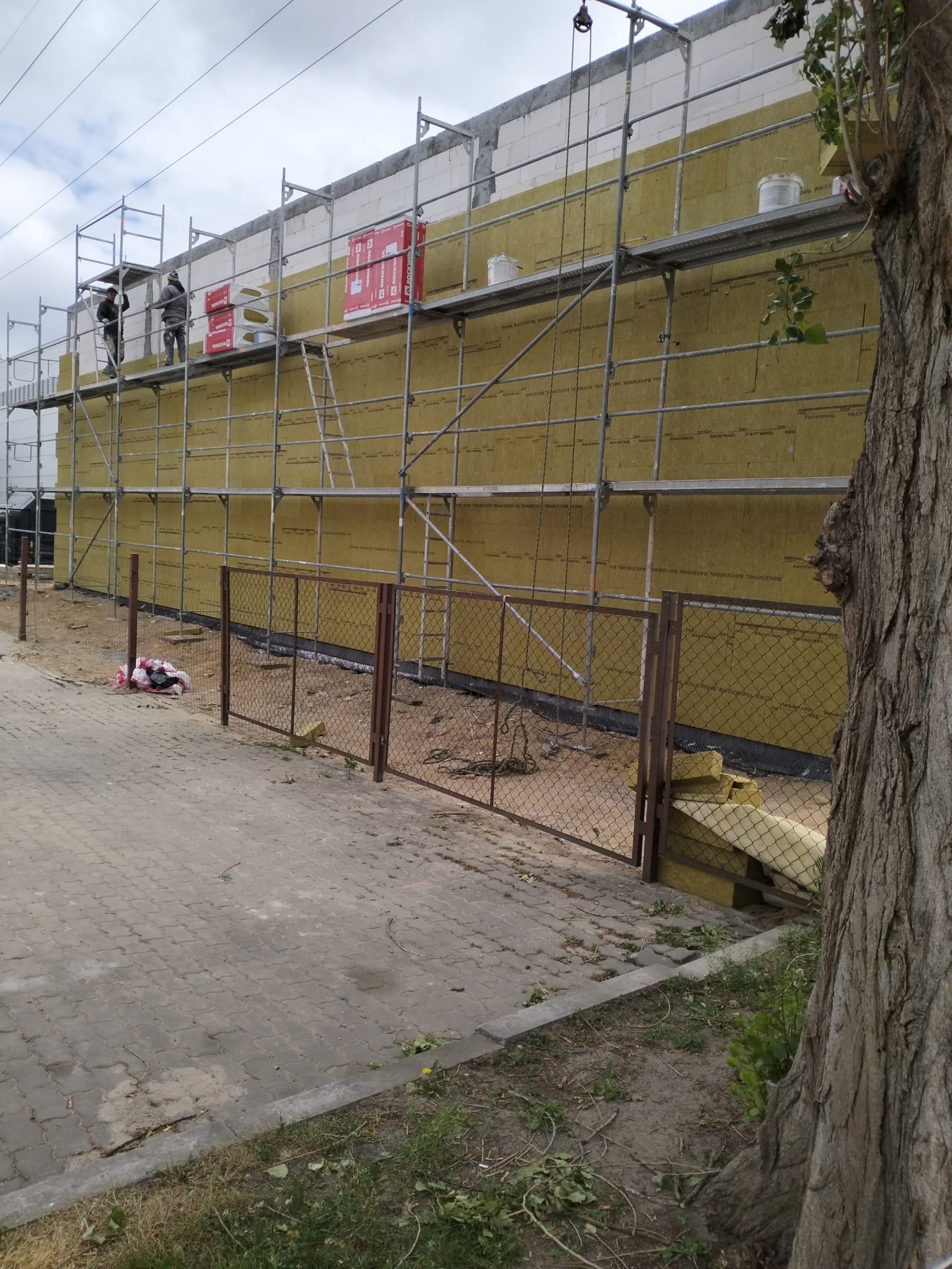 Dwuosobowa ekipa na rusztowaniu montuje żółtą izolację na ścianie budynku z białych bloczków, widoczna brama z siatki i fragment pnia drzewa.