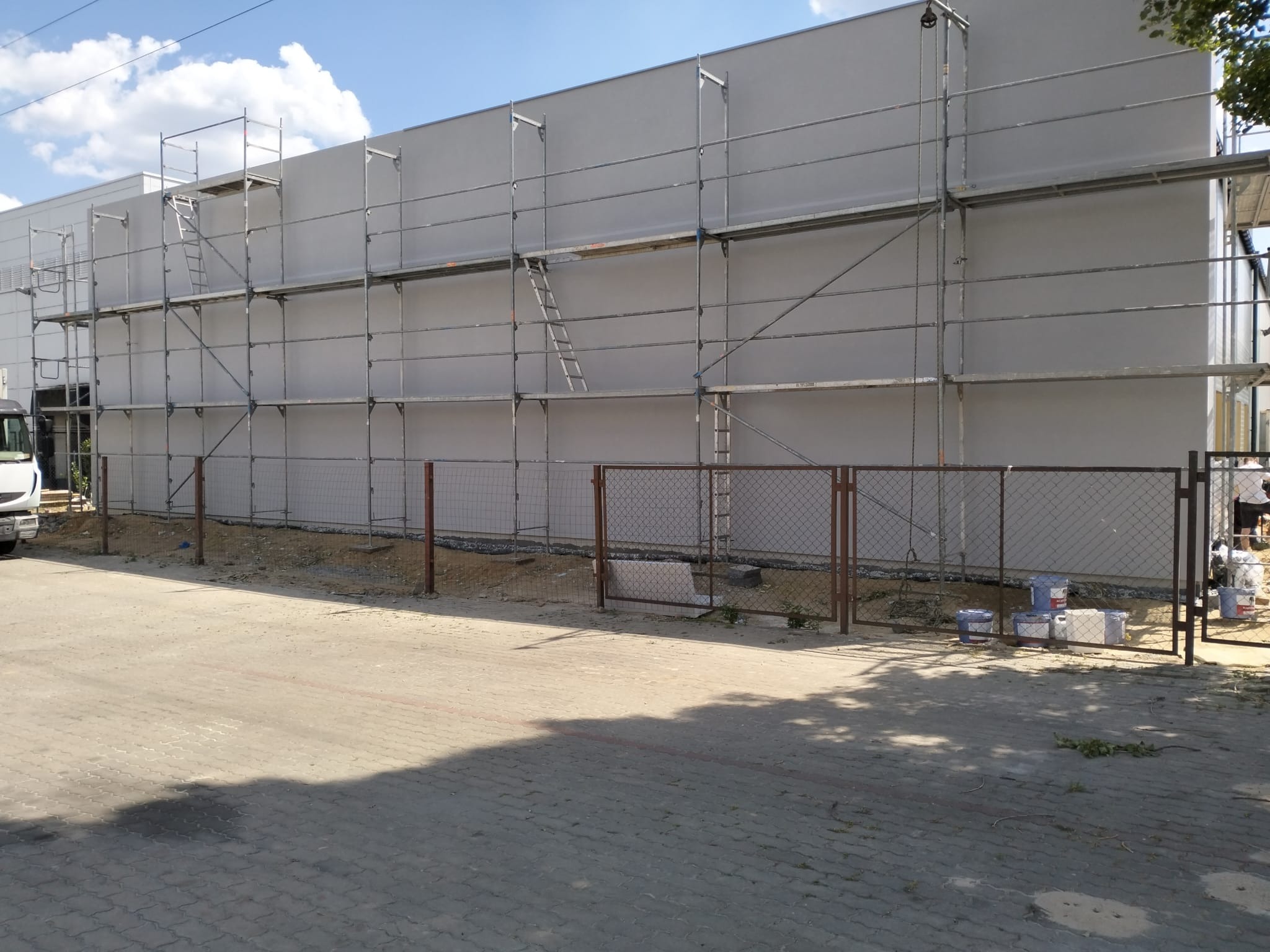 Budynek przemysłowy w trakcie prac elewacyjnych, widoczne rusztowania na całej ścianie, puste wiadra po farbie i siatka ogrodzeniowa.