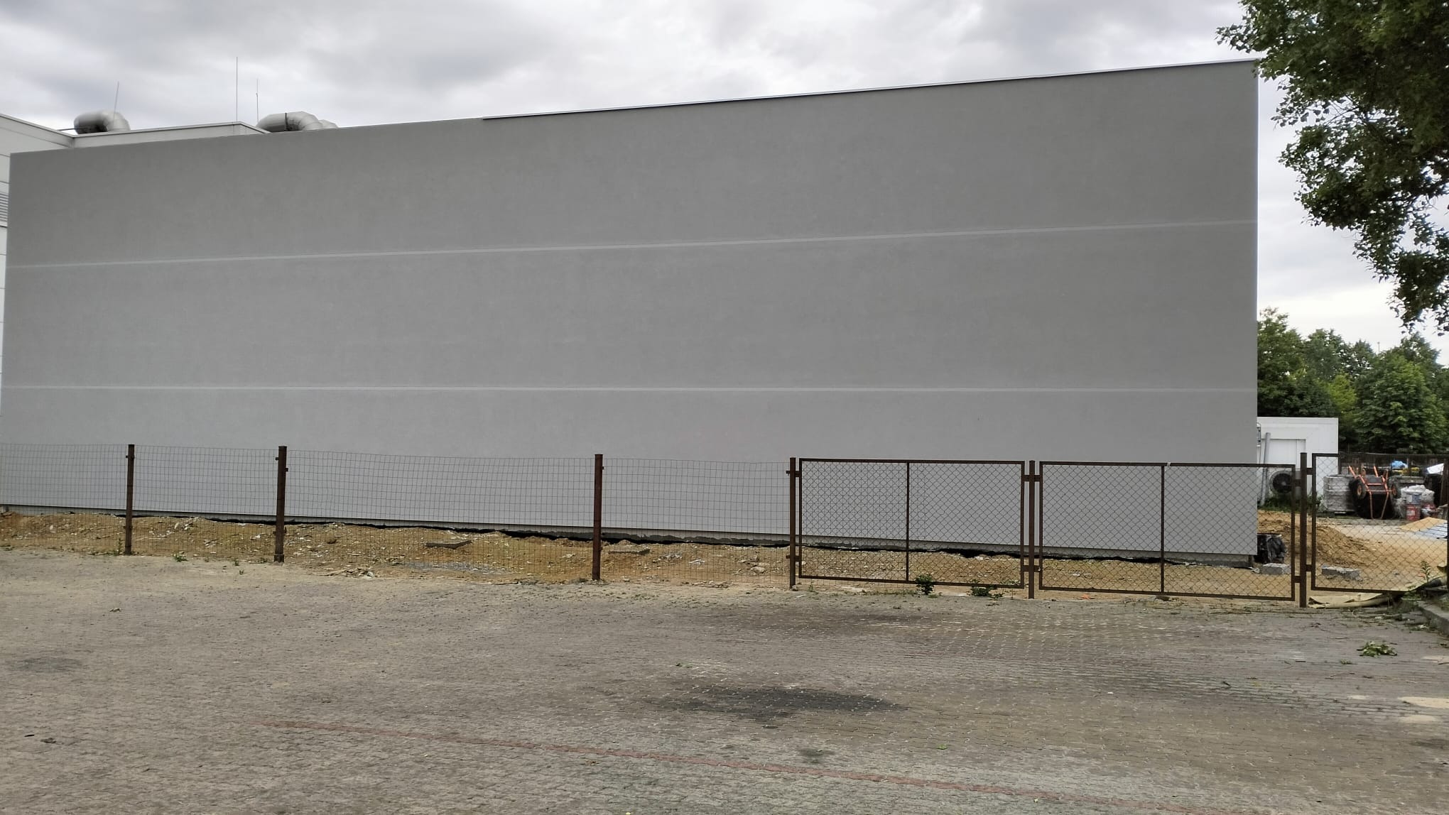 Szara, gładka elewacja budynku z widocznymi poziomymi pasami, ogrodzona siatką z brązowymi słupkami, na tle pochmurnego nieba i zieleni drzew.