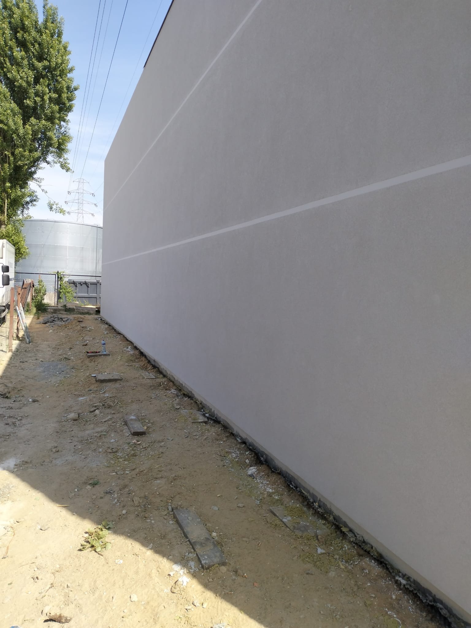 Gładka, jasnoszara elewacja budynku z widocznymi poziomymi pasami, teren wokół w trakcie prac wykończeniowych, widoczny zbiornik przemysłowy w tle.