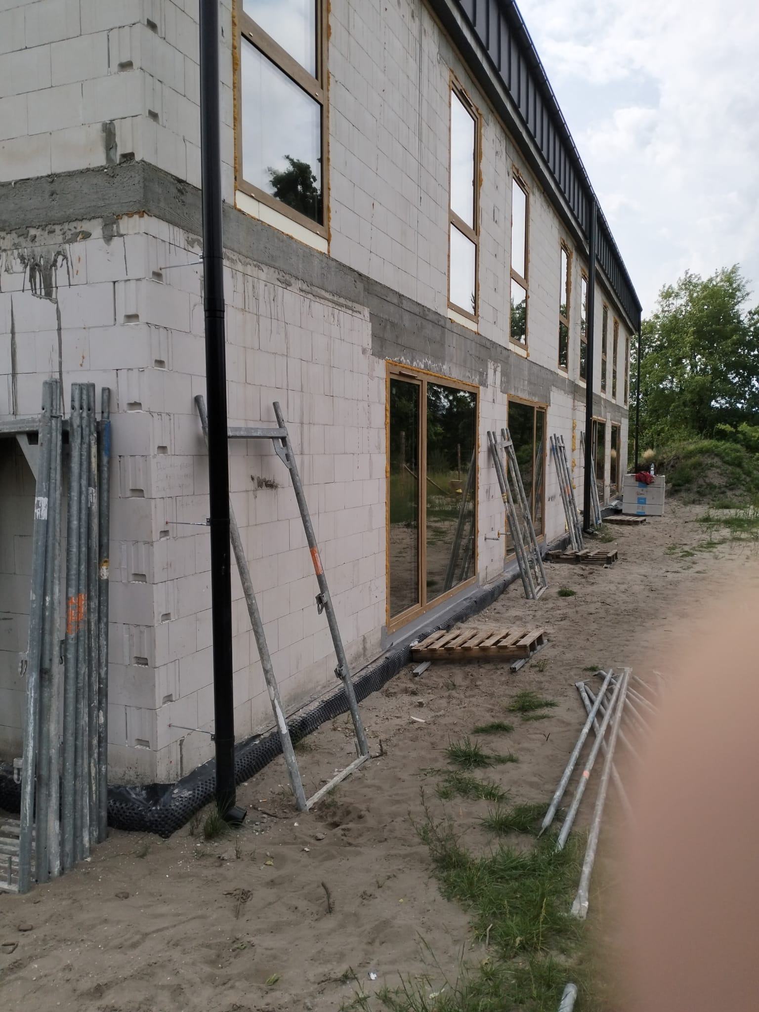 Budynek w trakcie budowy, ściany z białego bloczka, wstawione okna z drewnianą ramą, widoczne rusztowania i drabiny opierające się o ścianę, grunt wokół budynku piaszczysty.