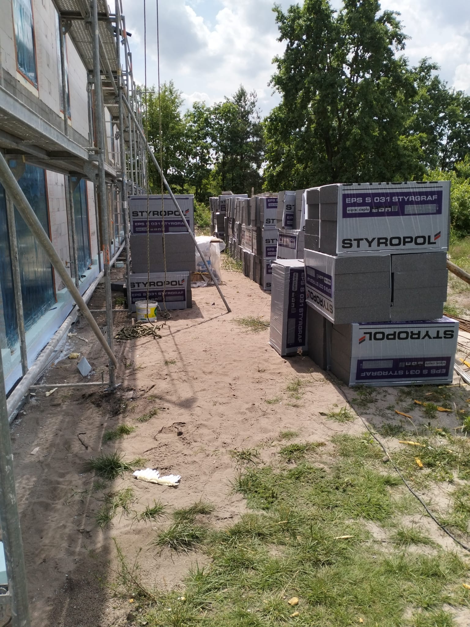 Styropian grafitowy ułożony w stosy obok budynku w trakcie budowy, widoczne rusztowania i przygotowane ściany.