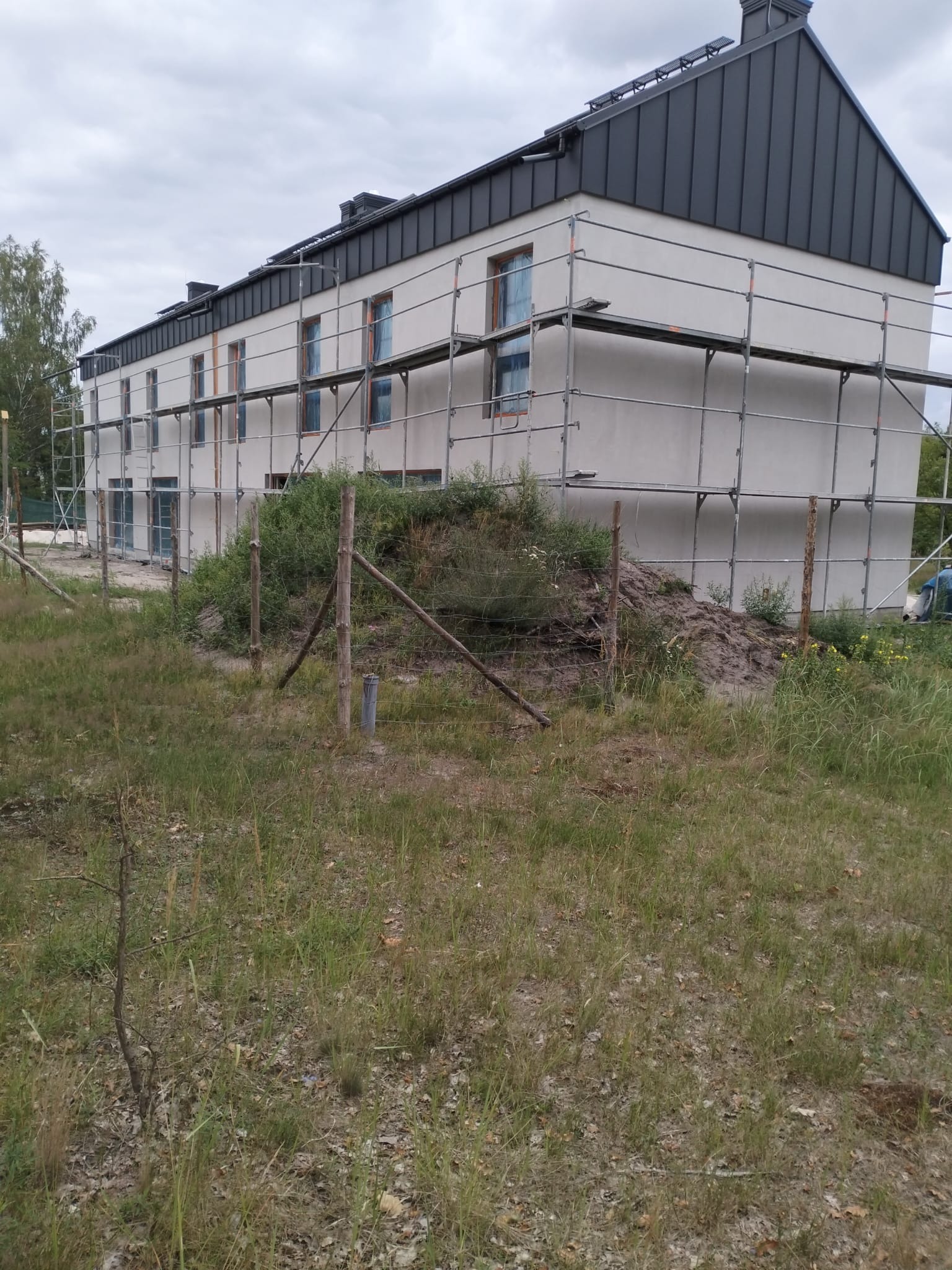 Budynek w trakcie ocieplania elewacji, widoczne rusztowania i częściowo otynkowana ściana, ogrodzenie z drewnianych słupków i siatki, zaniedbany teren wokół.