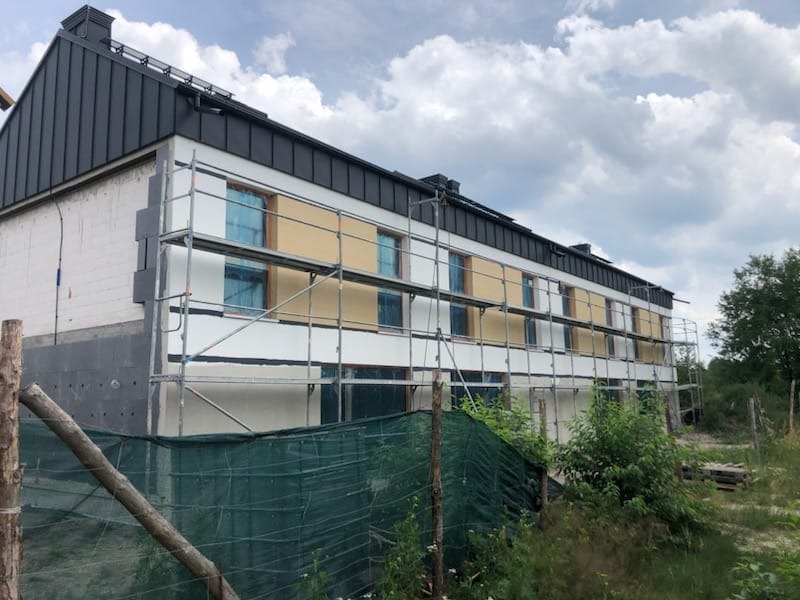 Dwupiętrowy budynek w trakcie prac elewacyjnych, widoczne rusztowania, częściowo ocieplona ściana z szarymi płytami izolacyjnymi, okna zabezpieczone folią, czarny dach i zielone otoczenie.