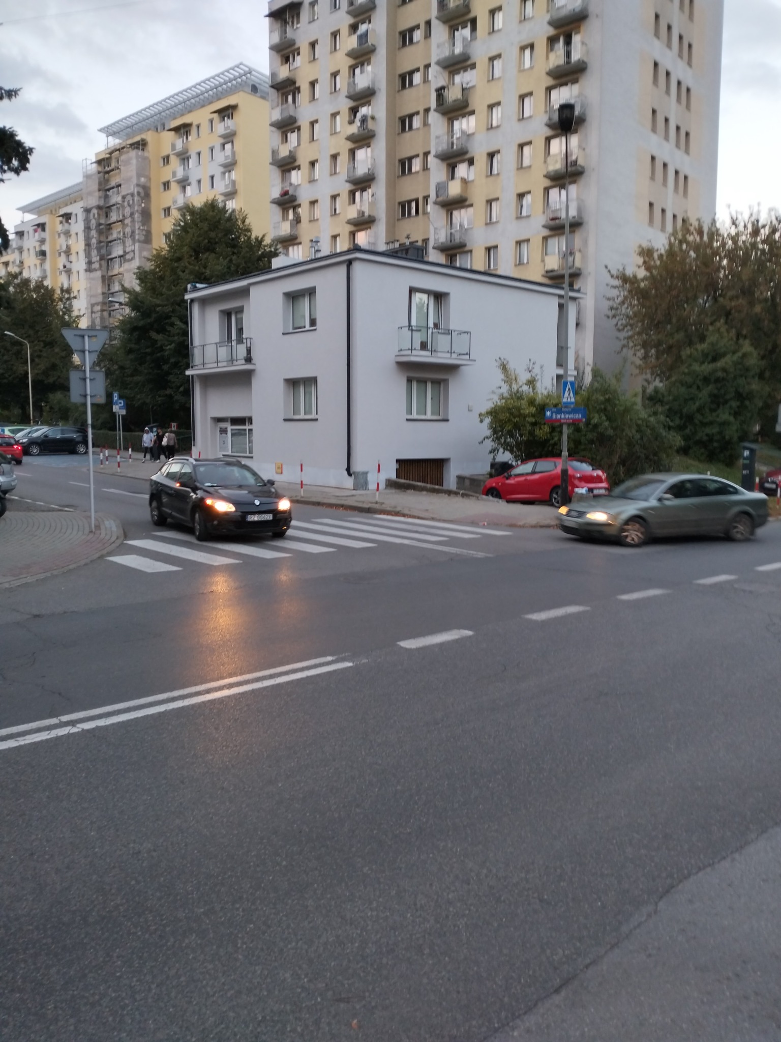 Dwupiętrowy budynek mieszkalny z białymi ścianami i balkonami, widoczny z ulicy z przejściem dla pieszych, na tle wysokich bloków mieszkalnych.