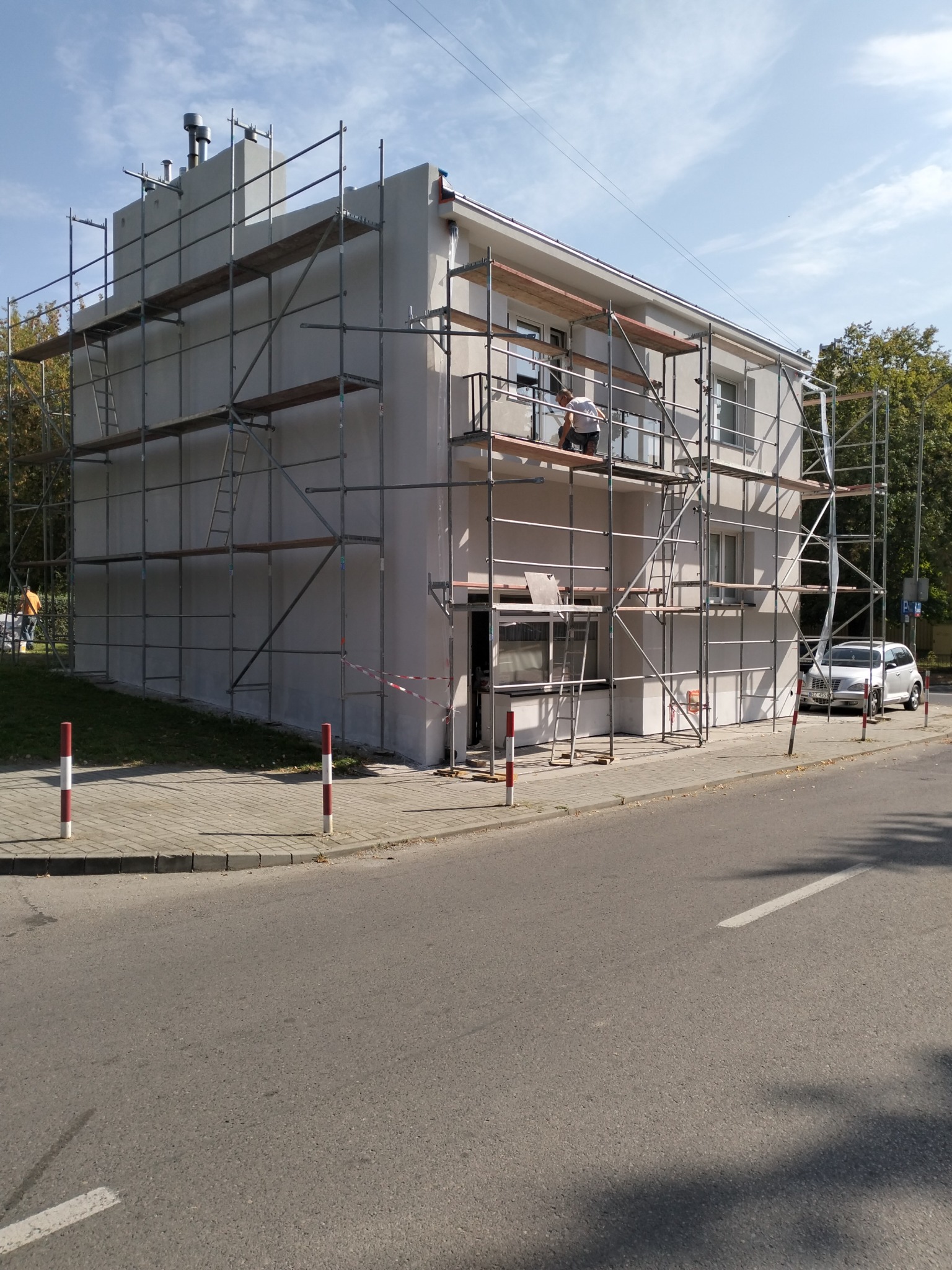 Dwupiętrowy budynek w trakcie prac elewacyjnych, otoczony rusztowaniem, z widocznym pracownikiem na rusztowaniu, częściowo otynkowana fasada, zaparkowany samochód osobowy w tle.