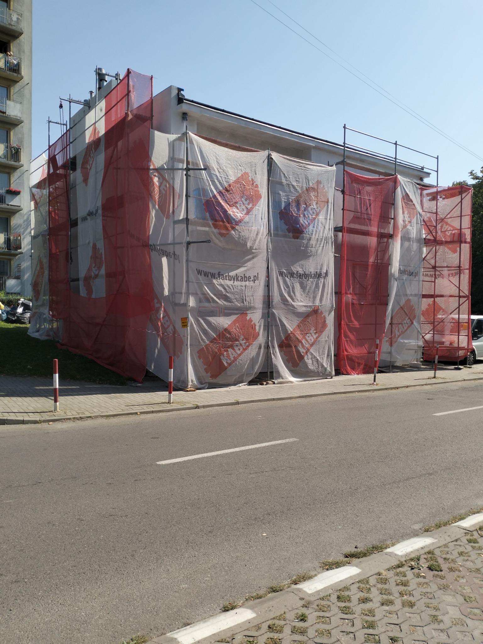 Budynek w trakcie prac elewacyjnych, osłonięty rusztowaniami i siatką ochronną z nadrukiem reklamowym, widok z poziomu ulicy.